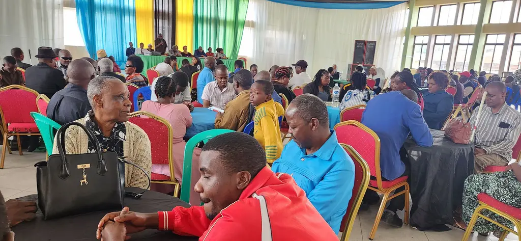 Delegates at a TLB general assembly seated at tables, several with white canes resting beside them, listening to a speaker at the platform.