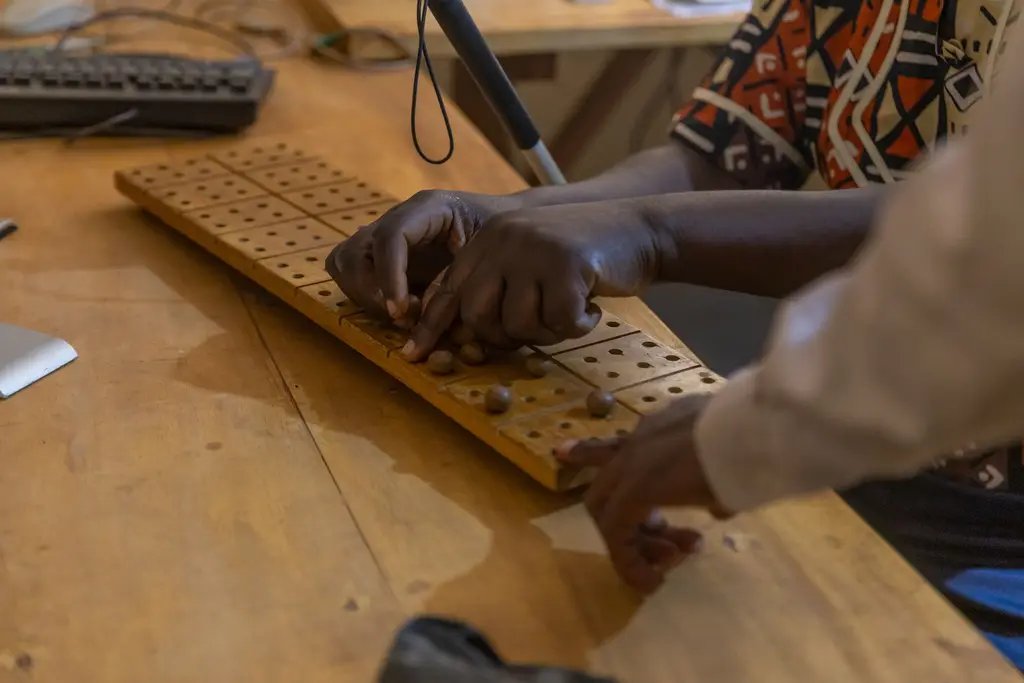 Close view of two pairs of hands at a wooden Braille teaching board, setting round pegs into drilled holes to build Braille cells. A keyboard and a folded white cane lie on the desk behind.