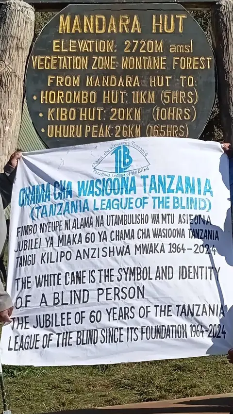 Climbers hold a TLB banner at the Mandara Hut sign, 2,720 metres on Kilimanjaro. The banner reads “The white cane is the symbol and identity of a blind person” and marks 60 years of the Tanzania League of the Blind, 1964–2024.