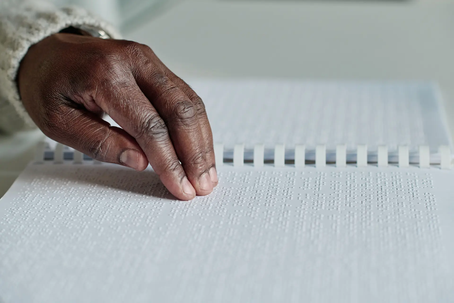 A reader's hand moves across a line of Braille in an open, spiral-bound Braille book.